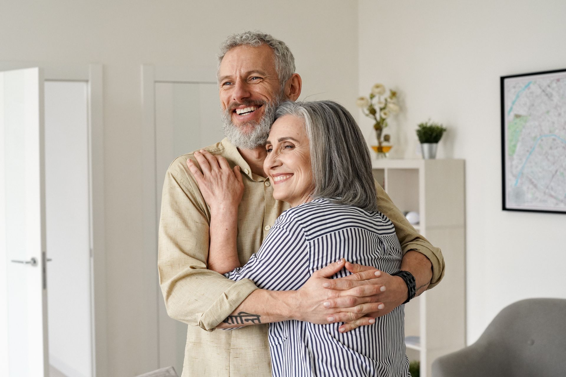 Couple embracing indoors, smiling. Man in tan shirt, woman in striped shirt. Light-filled room with art.