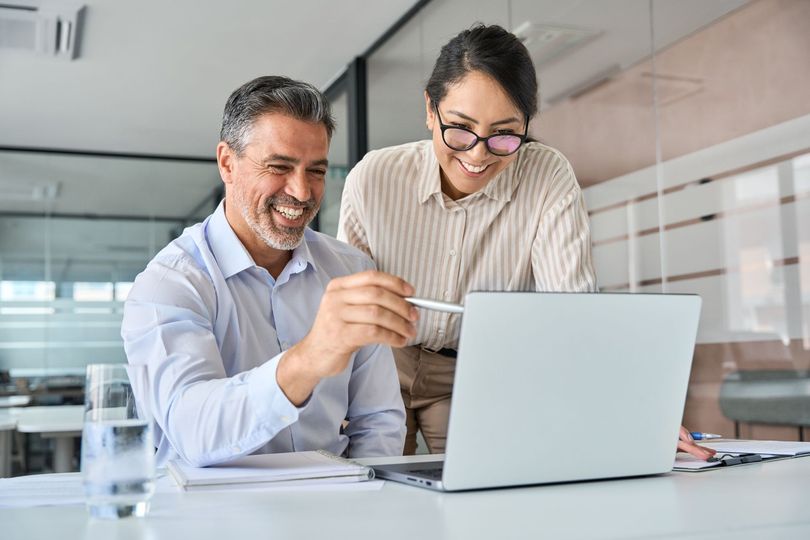 Two people looking at a laptop, smiling. Man points with a pen, woman leans over, office setting.