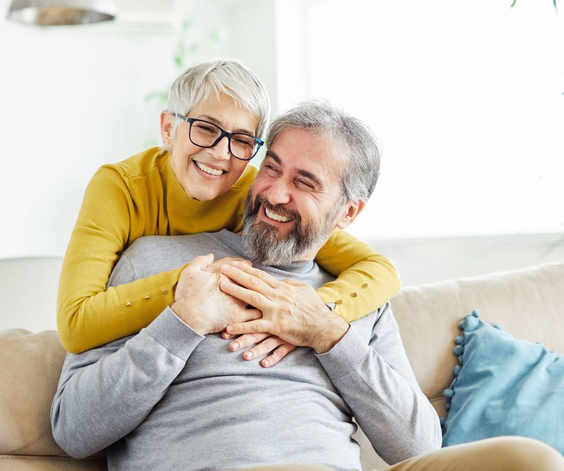 Smiling older couple embracing on a couch, woman with glasses, man with beard, cozy home setting.