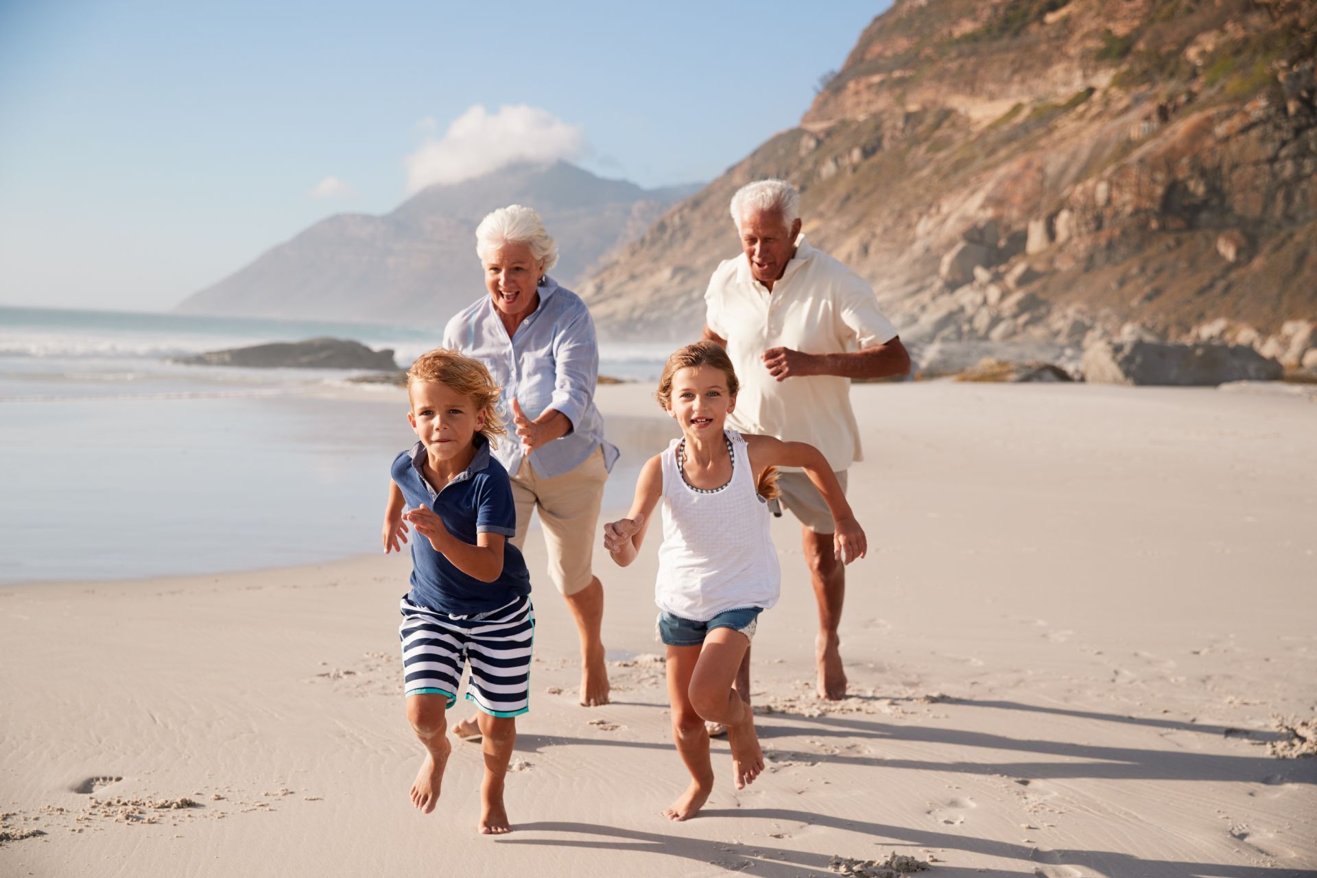 Grandparents and grandchildren running on a sandy beach near the ocean. Mountains in background.
