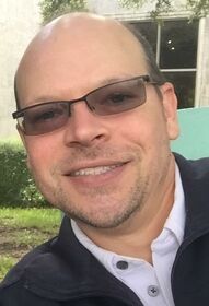 Man with glasses smiling, wearing a dark jacket, outdoors.