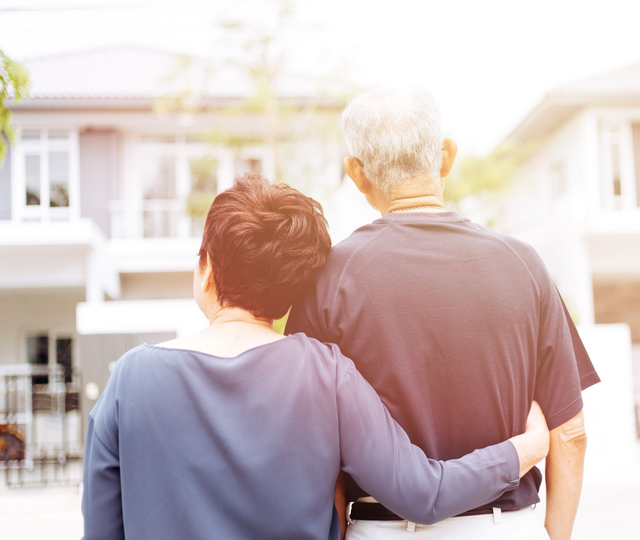 Elderly couple with arms around each other looking towards a house in the sunlight.