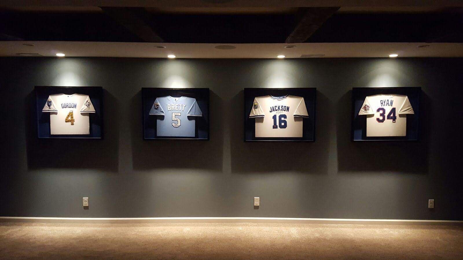Four framed jerseys with numbers 4, 5, 16, and 34 are displayed on a dark grey wall under recessed ceiling lights.