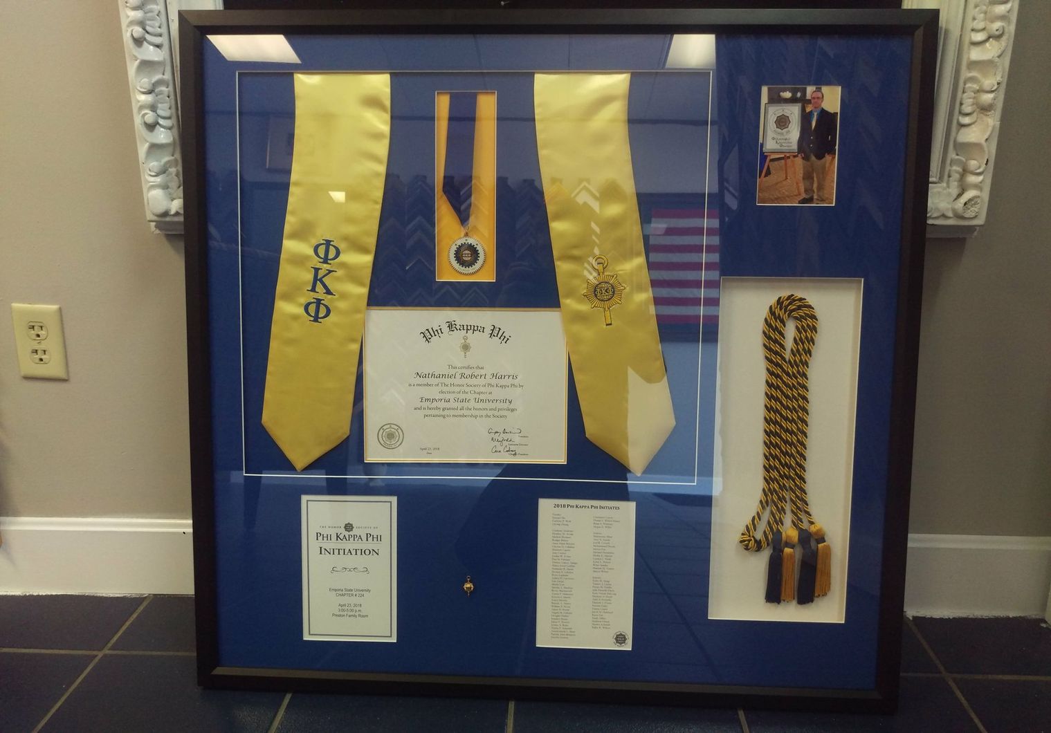 A framed display containing a gold Phi Kappa Phi graduation stole, certificate, honor cord, a medal, and a small photo.