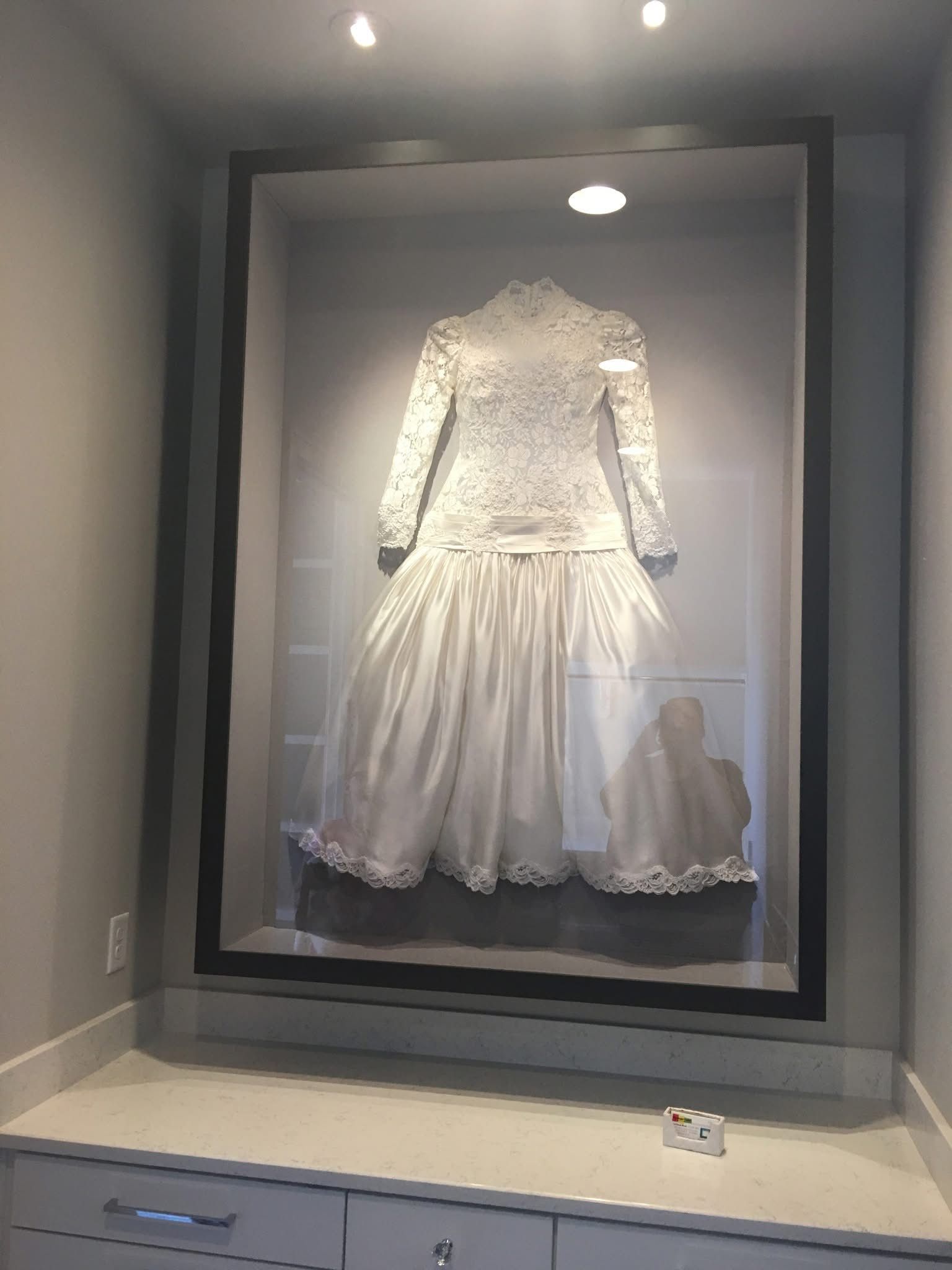 A white lace and satin wedding gown displayed behind glass in a wall-mounted case above a white counter.