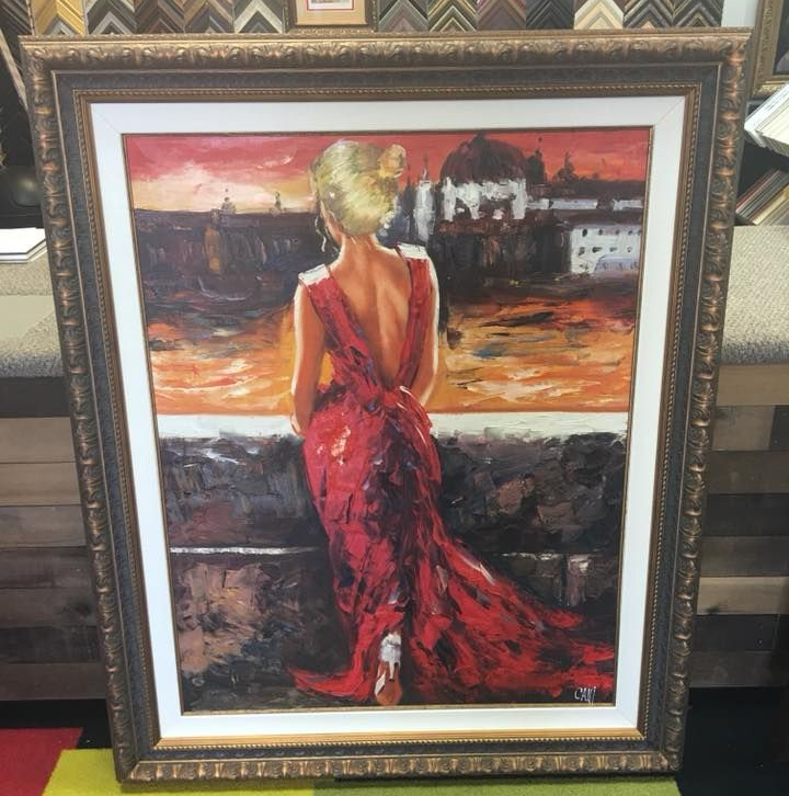 A framed painting of a person in a red gown standing with their back to the viewer, looking out over a city at sunset.