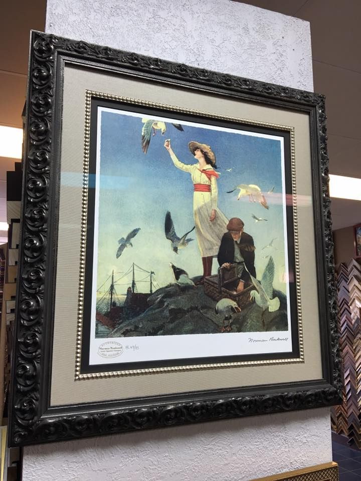 A framed print shows a person feeding birds on a rocky outcrop near a body of water with a bridge in the distance.