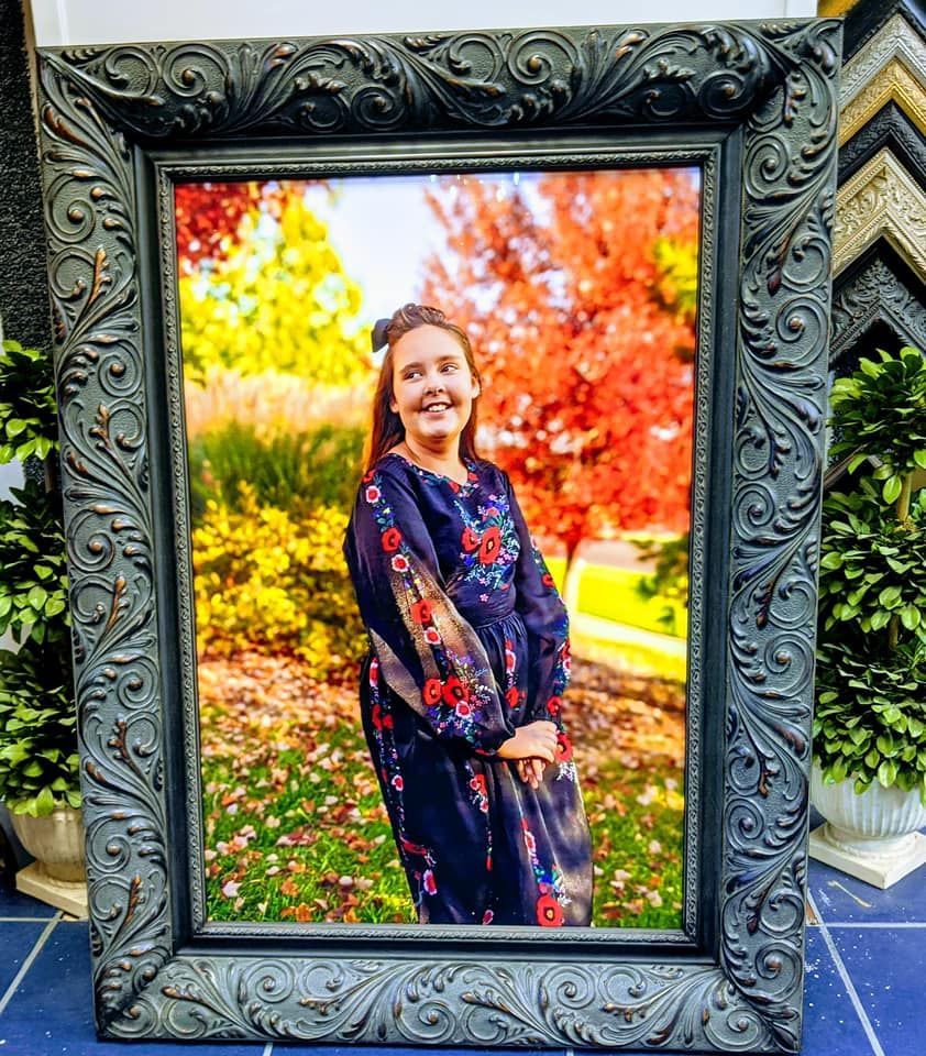 A person in a floral, dark-colored long-sleeve dress poses in an autumn landscape, framed in an ornate, dark gray frame.