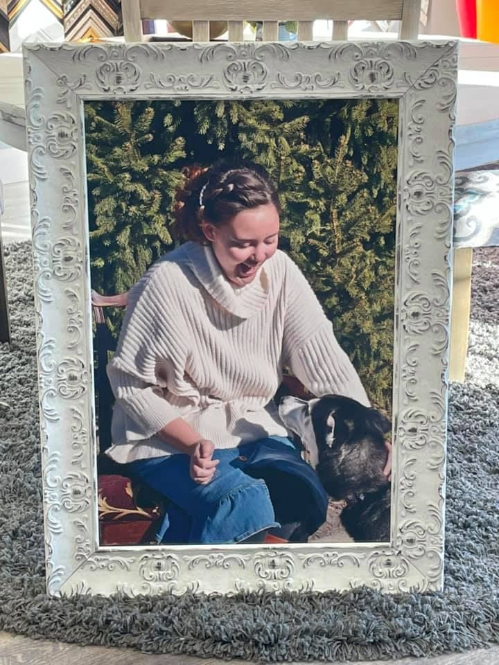 A person in a cream cowl-neck sweater and blue pants smiles while petting a dark-furred dog in a vintage white frame.