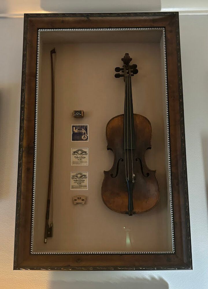 A wooden shadow box displays a vintage violin, a bow, a bridge, and small labels mounted on a light-colored background.