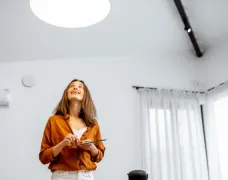 A woman is standing in a living room holding a tablet and looking up at the ceiling.