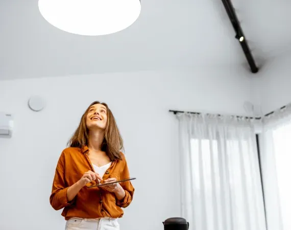 A woman is standing in a living room holding a tablet and looking up at the ceiling.
