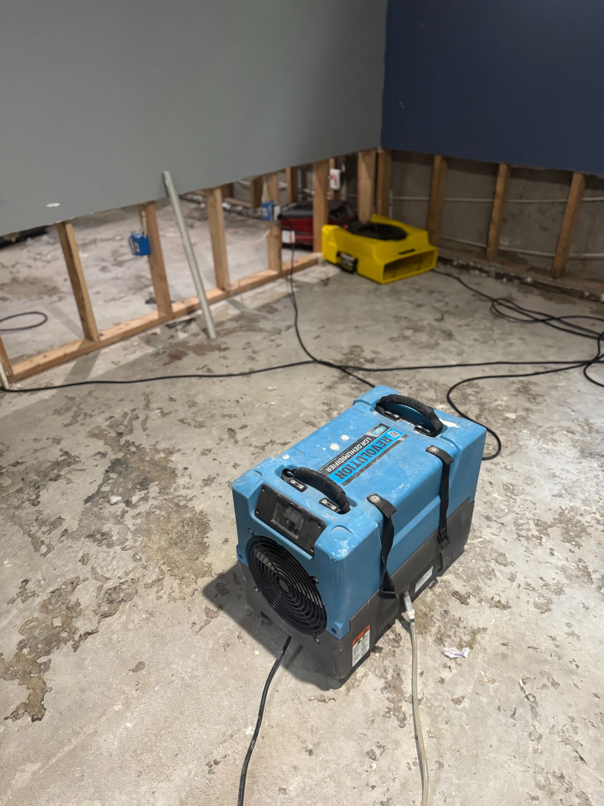 Flood-damaged room with two portable dehumidifiers, exposed wall studs, and wet concrete floor