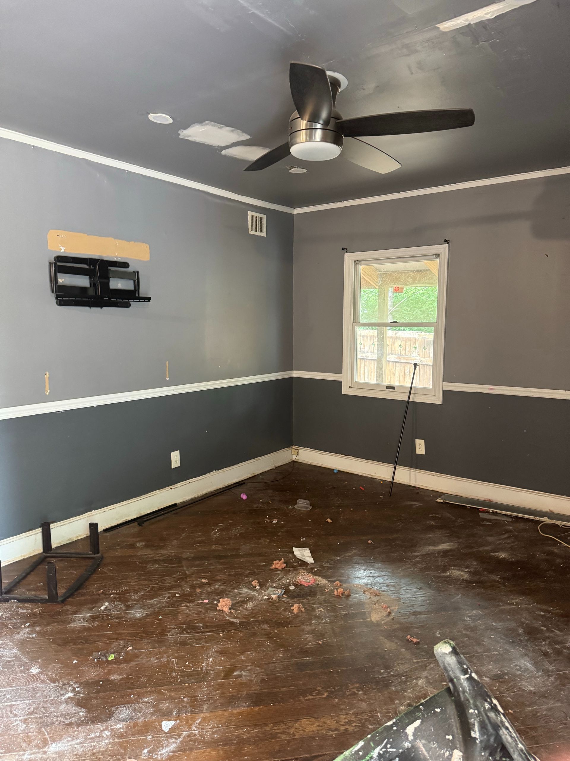 Empty gray room with damaged dark floor, ceiling fan, window, and wall-mounted TV bracket.