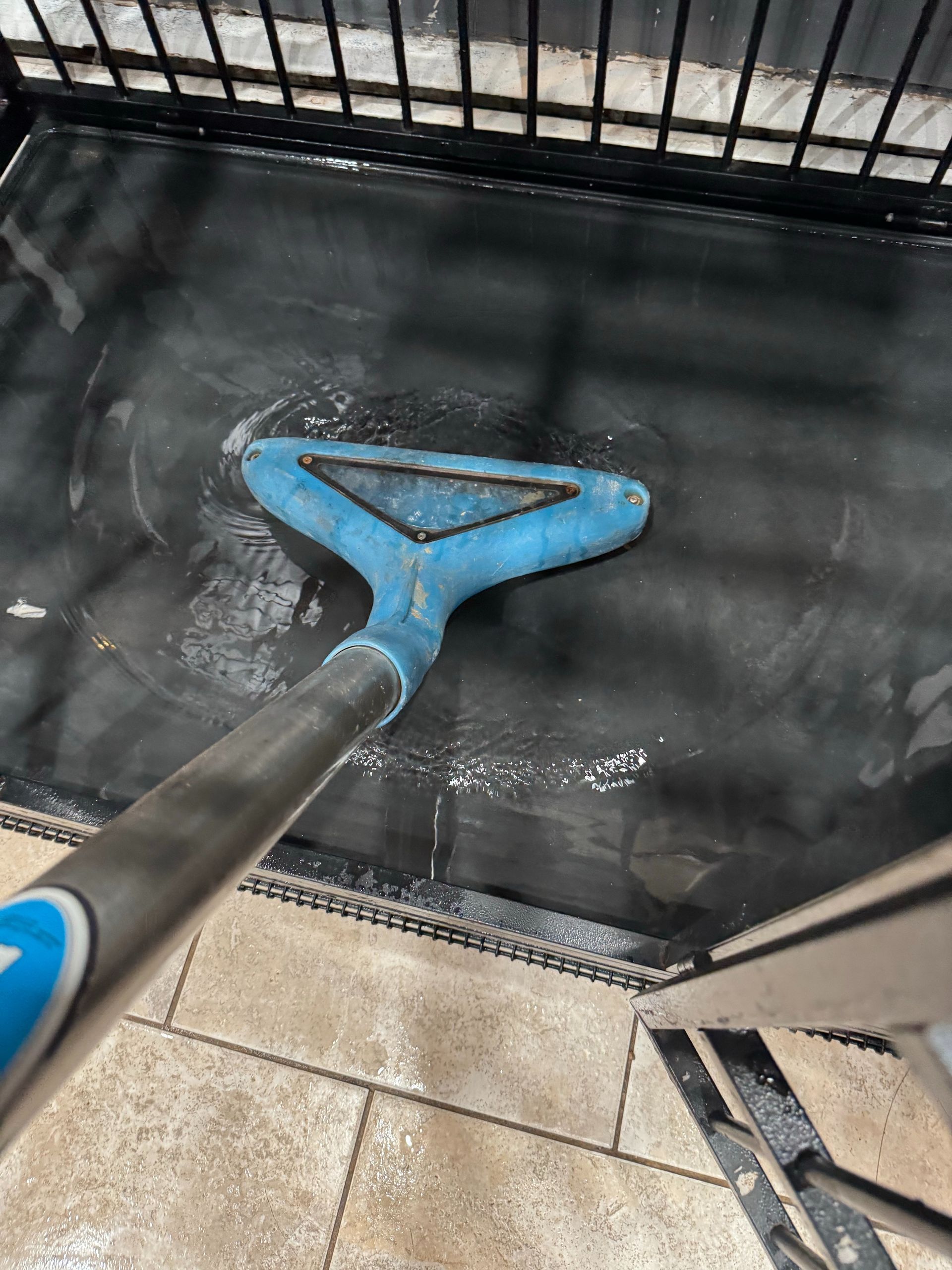 Blue floor scrubber cleaning a wet black surface near a metal fence and tiled floor