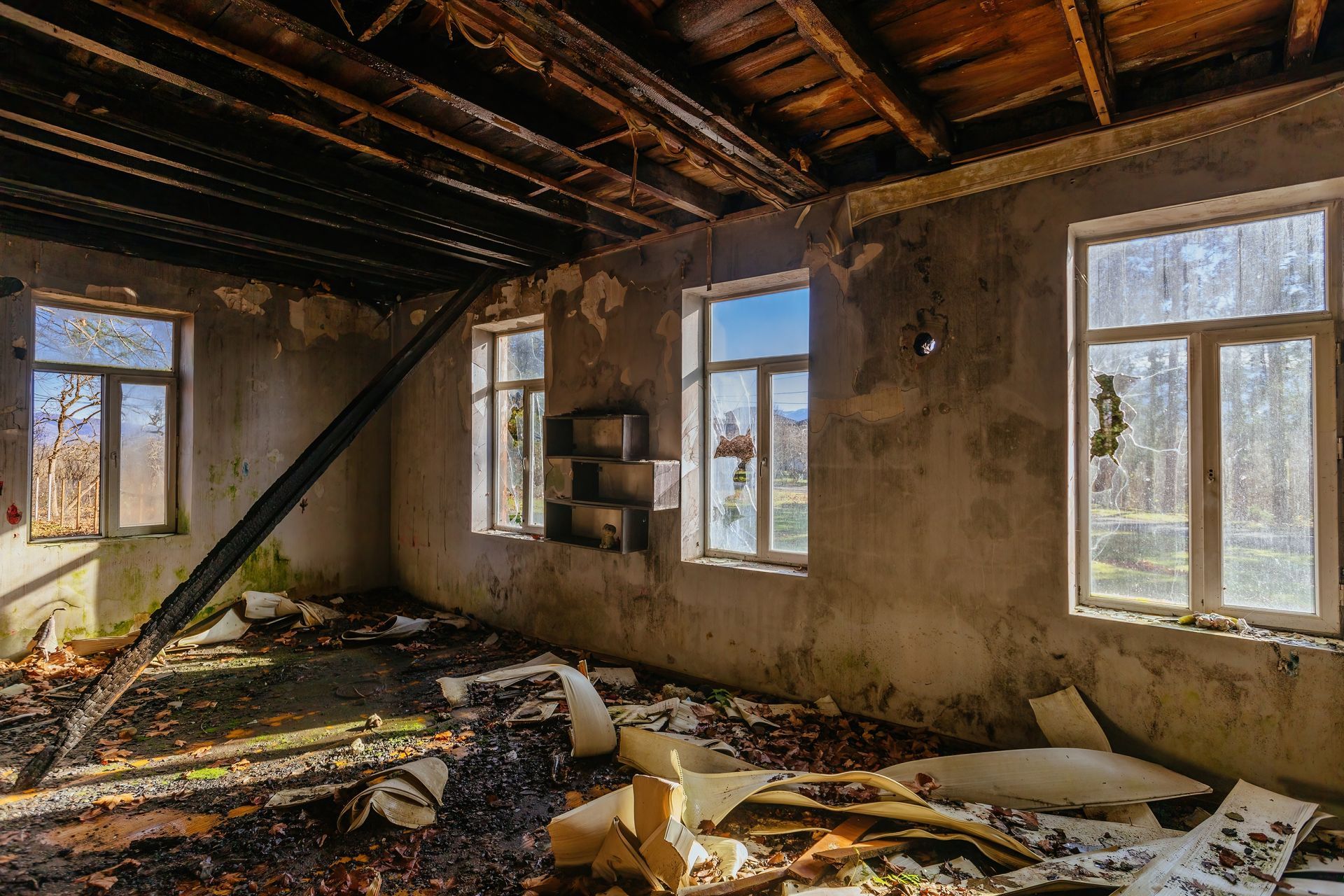 Abandoned room with debris on the floor, exposed ceiling beams, and sunlight through broken windows