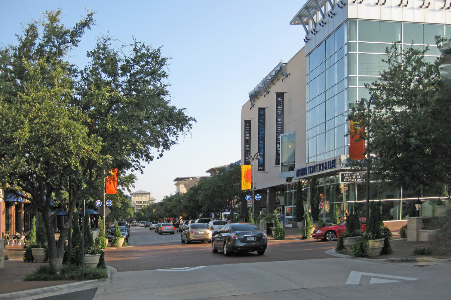 Tree-lined street with cars passing a modern glass-front building on a sunny day