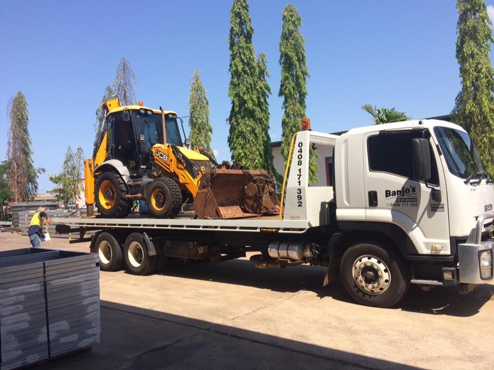 Yellow backhoe loader on a white flatbed truck, parked outdoors on a sunny day — Banjo's Anytime Towing in Humpty Doo, NT