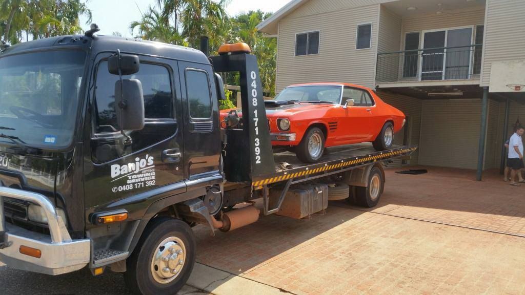 Black tow truck carrying an orange vintage car on a sunny day in front of a house — Banjo's Anytime Towing in Humpty Doo, NT