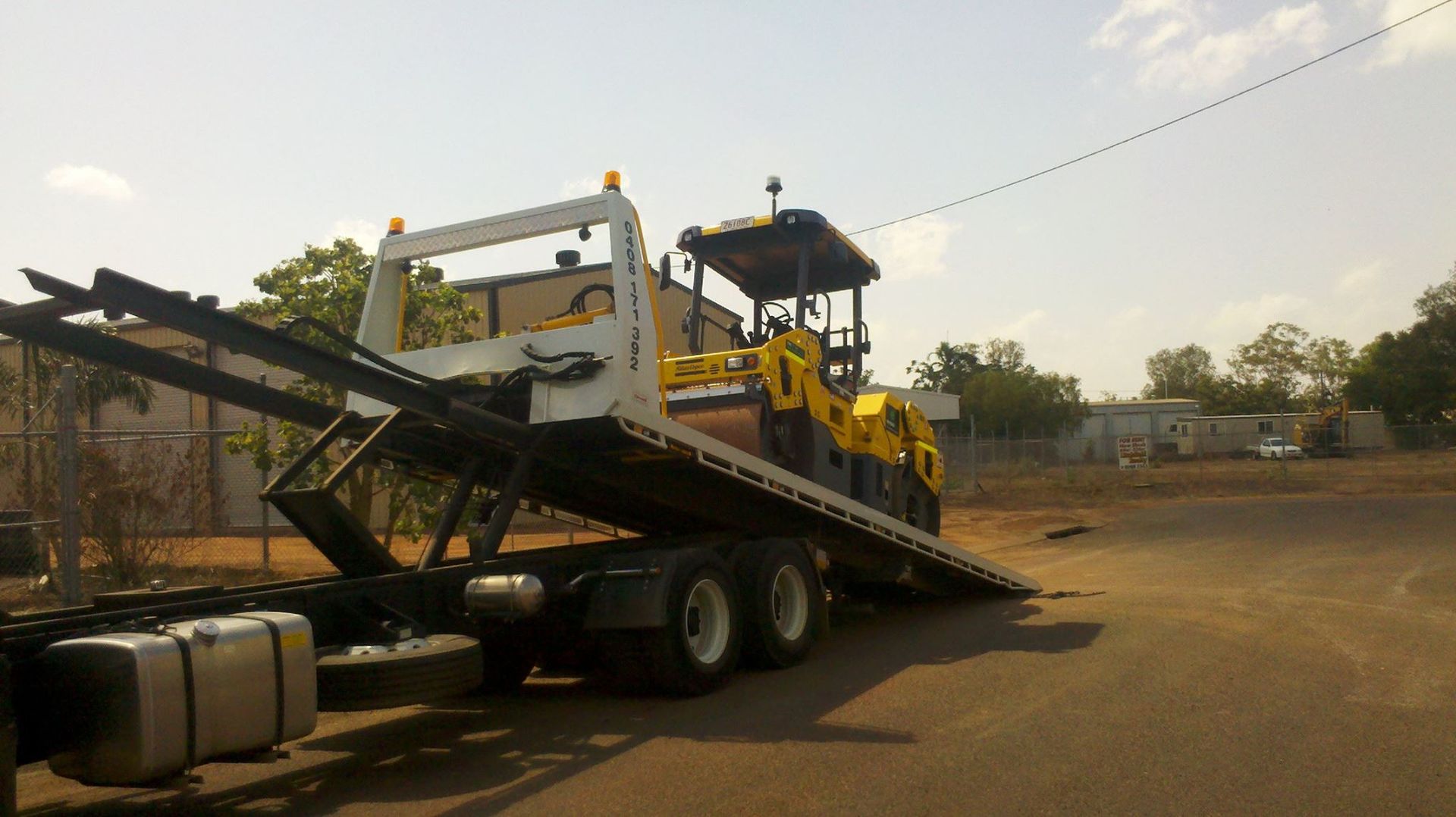 A yellow road roller is being loaded onto a flatbed truck on a sunny day — Banjo's Anytime Towing in Humpty Doo, NT