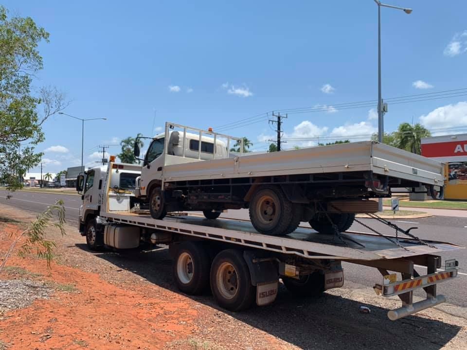 White flatbed tow truck carrying a light-coloured truck on a road with red dirt and blue sky — Banjo's Anytime Towing in Humpty Doo, NT
