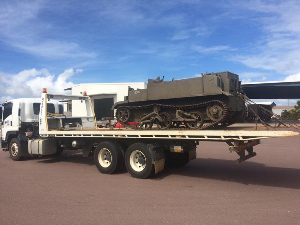 A flatbed truck carrying a tracked military vehicle on a tarmac under a blue sky — Banjo's Anytime Towing in Humpty Doo, NT