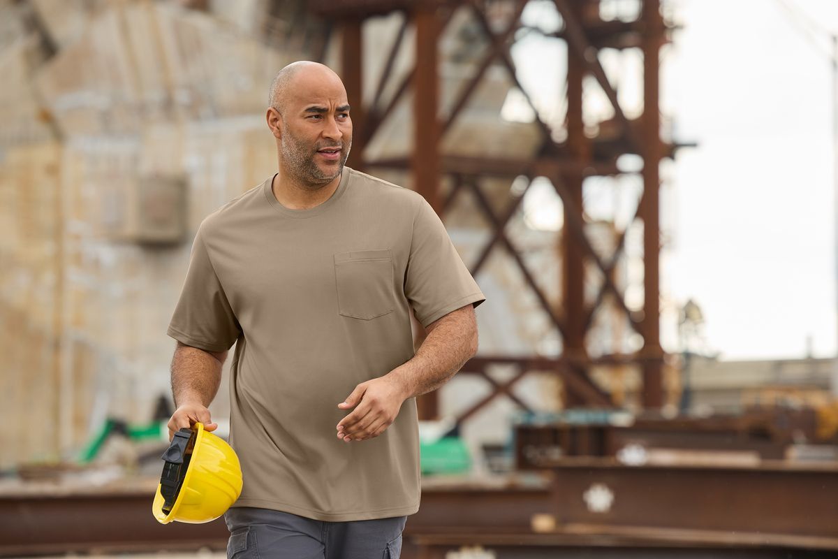 Man in work clothes holding a yellow hard hat, walking on a construction site.