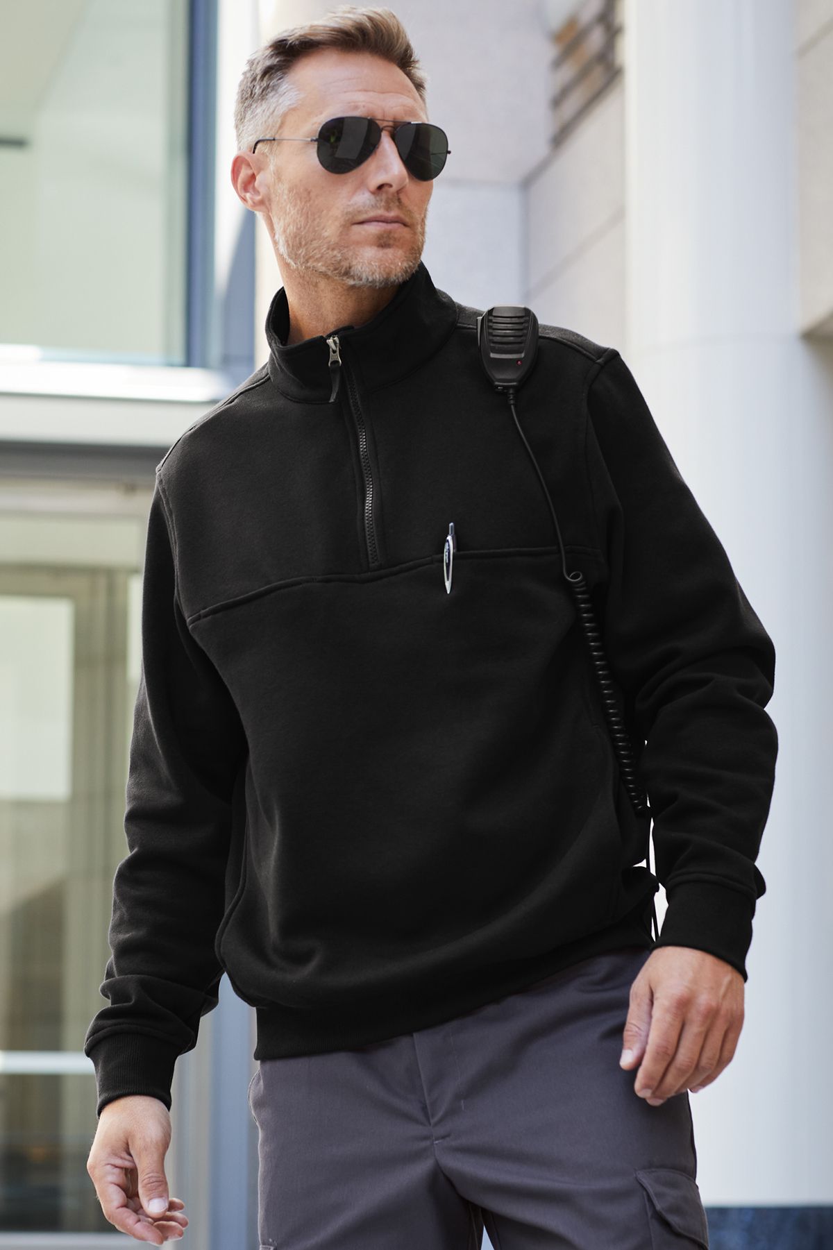Man in black sweatshirt, sunglasses, and cargo pants, holding a radio, standing outside a building.