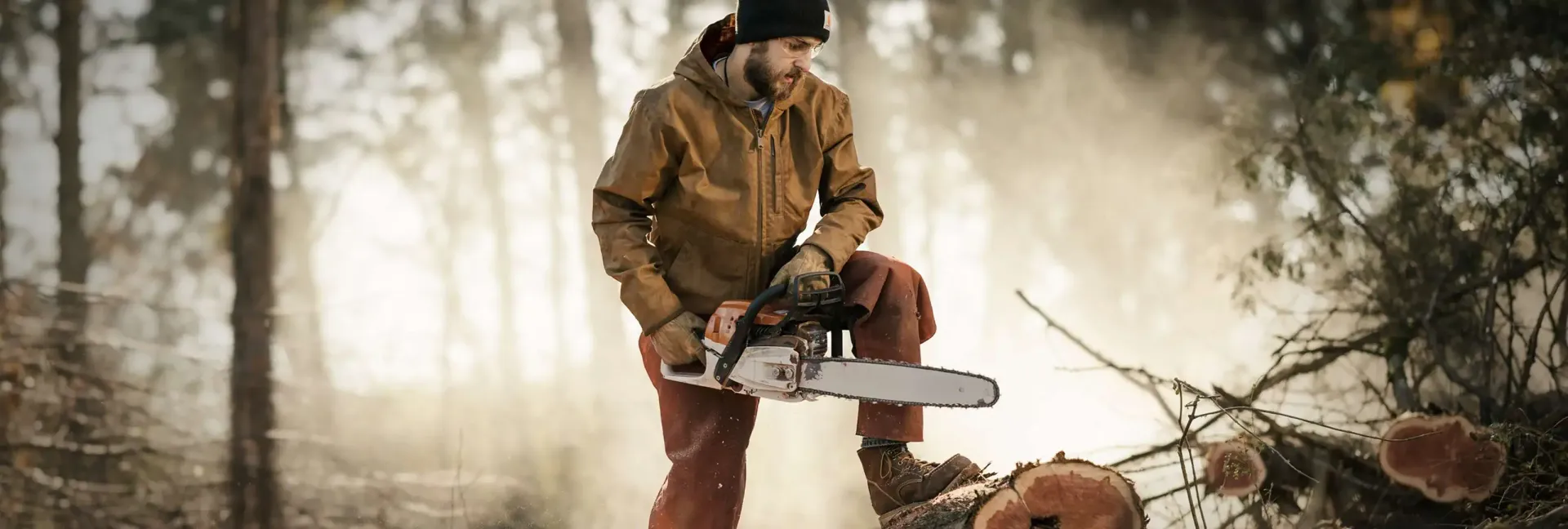 Man in brown jacket and hat with chainsaw, cutting wood in a forest.