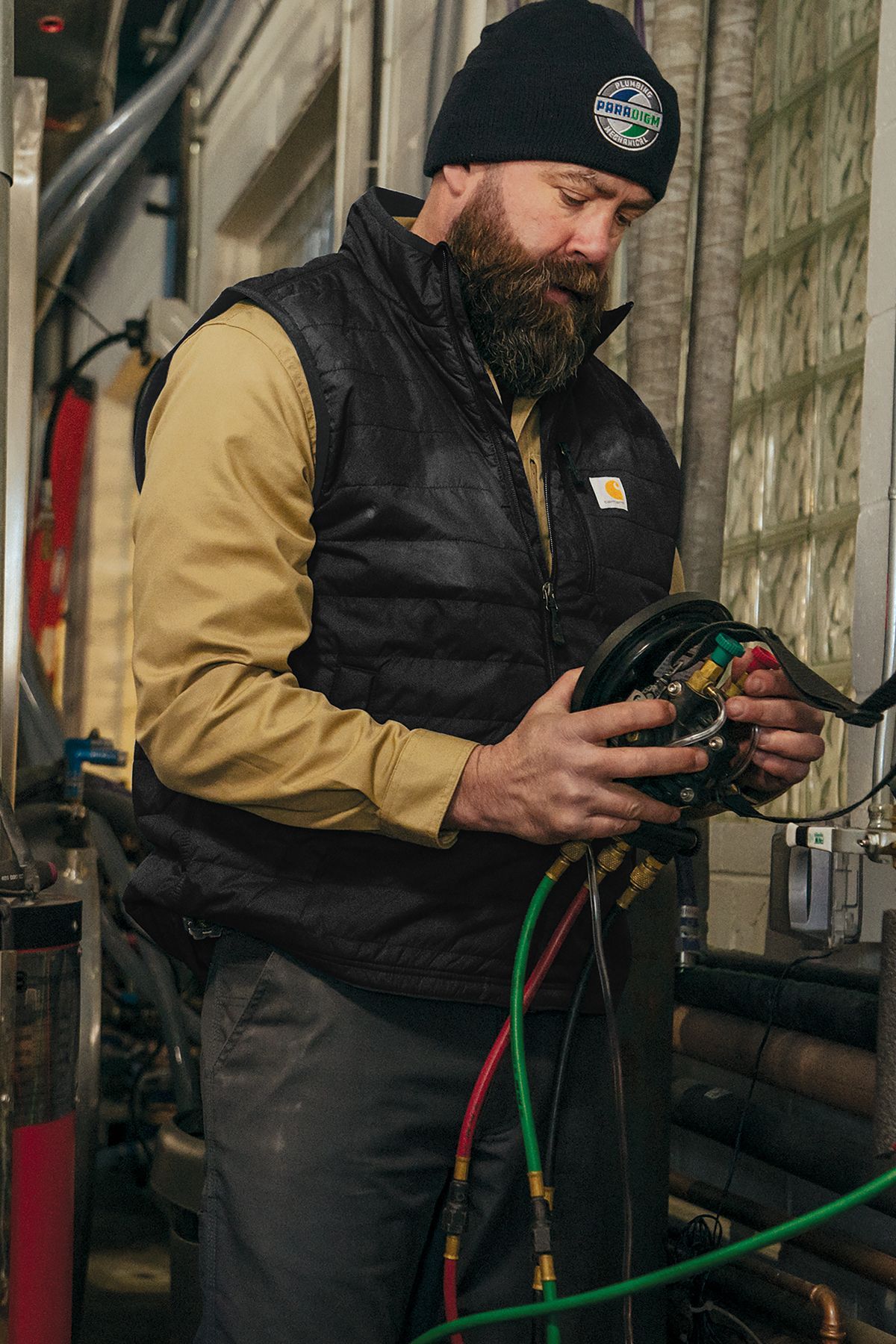 Bearded person in beanie and vest examines wires, inside a building.