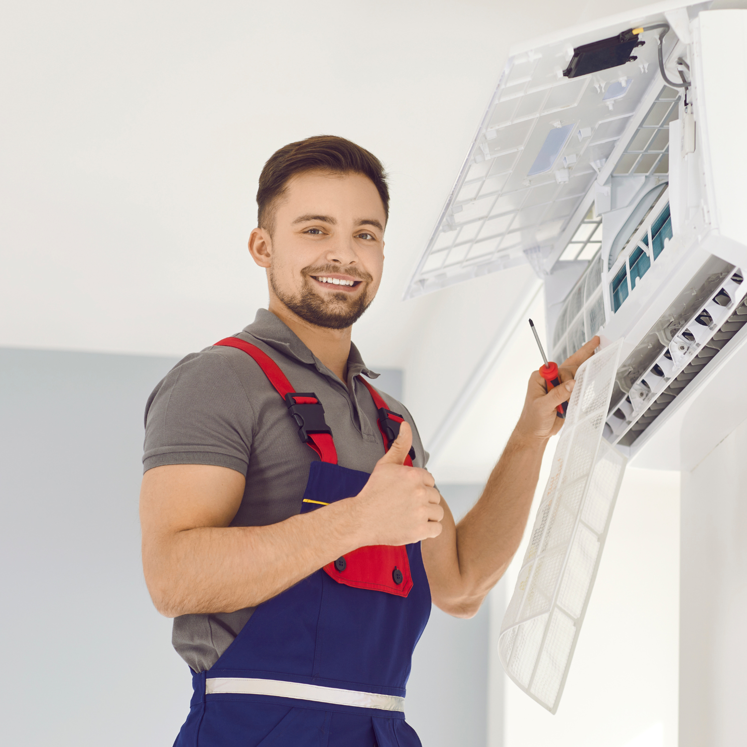 HVAC technician repairing an AC unit indoors, giving a thumbs up. Wearing overalls, smiling.
