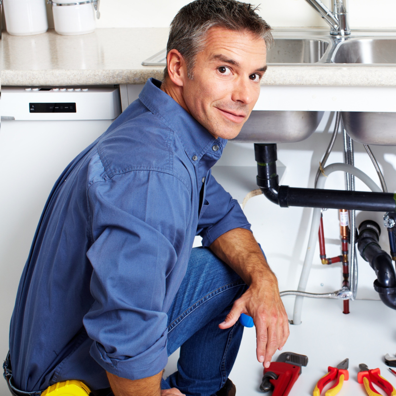 Plumber kneeling under a kitchen sink, fixing pipes with tools.