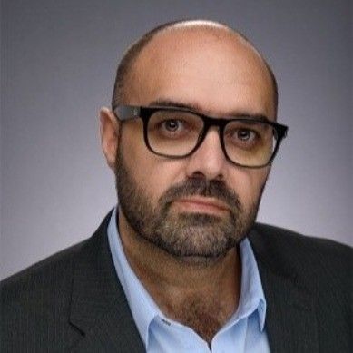A bald man with a beard and glasses is wearing a suit and blue shirt.