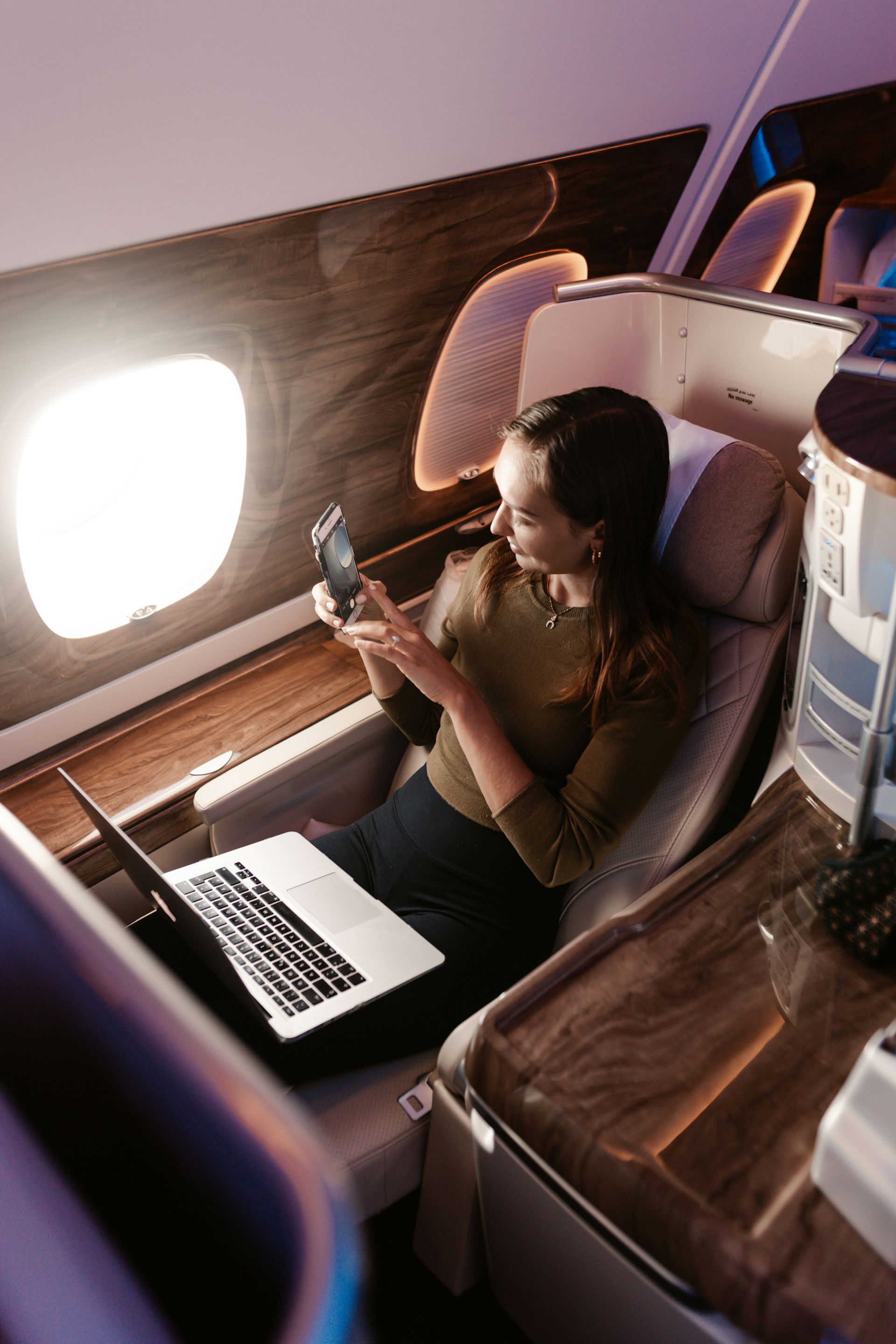 A woman is sitting on an airplane using a laptop and a cell phone.