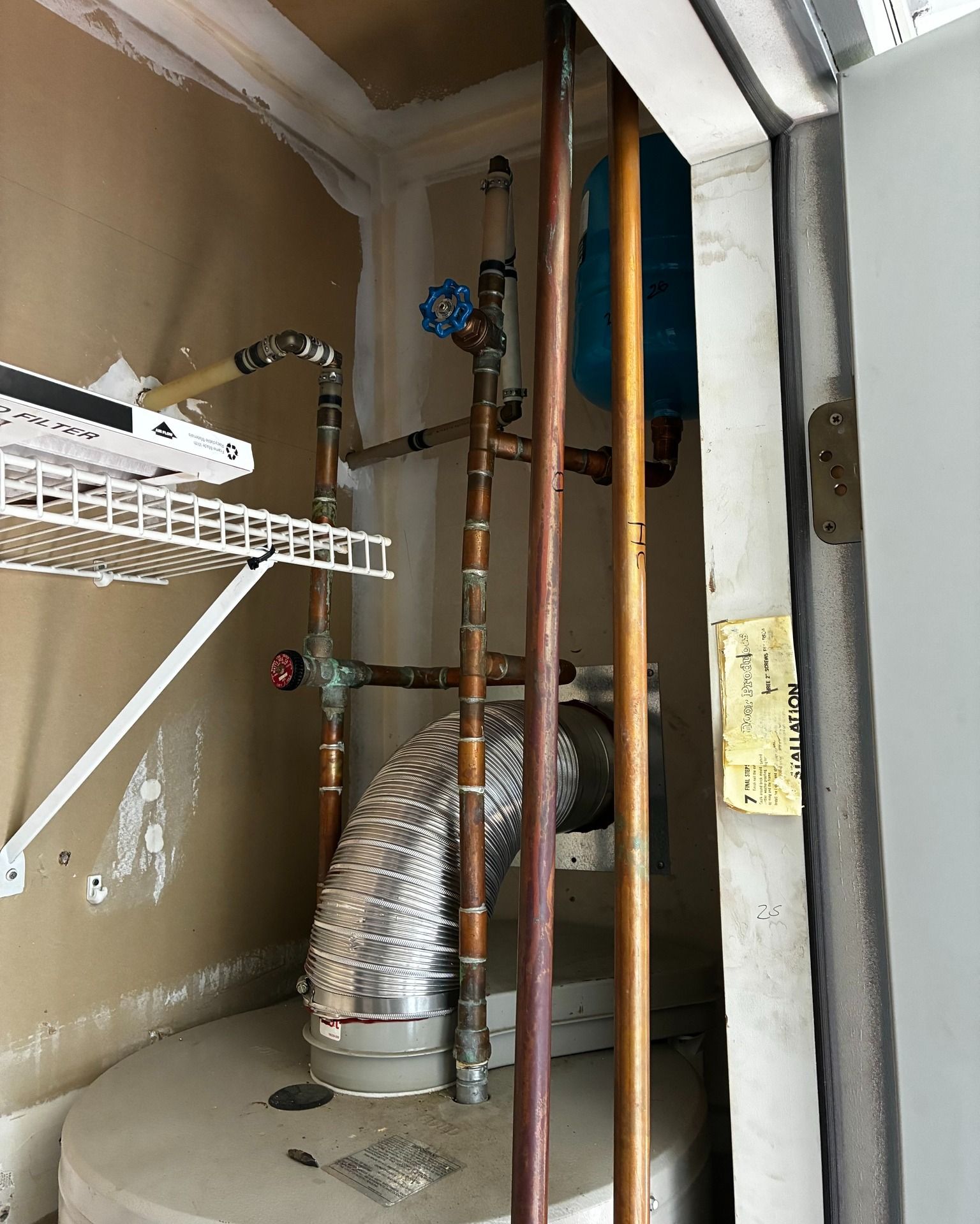 Copper pipes and a silver exhaust vent connected to a water heater inside a utility closet with a blue expansion tank.