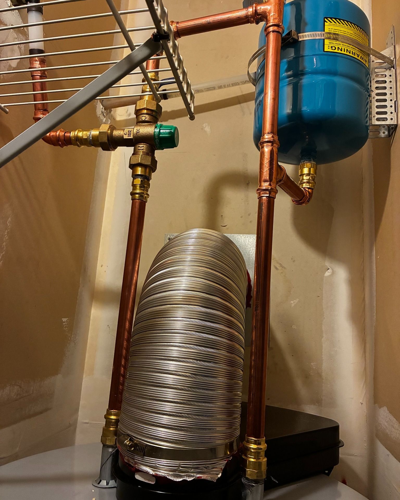 Copper pipes and a small blue expansion tank connected to a water heater unit, with a flexible metallic flue vent behind.