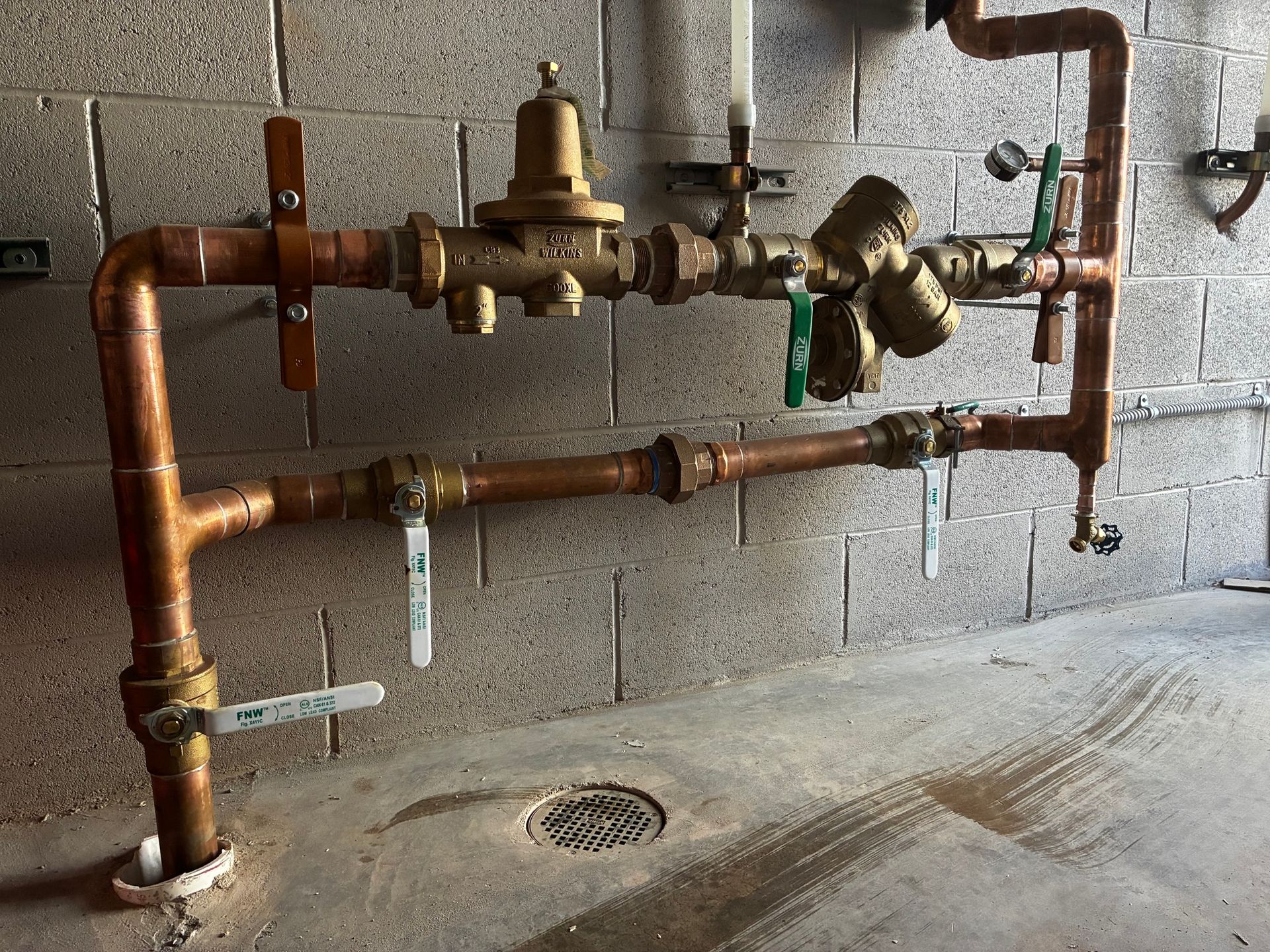 Copper piping system mounted on a block wall, featuring a pressure regulator, valves, and a Y-strainer.