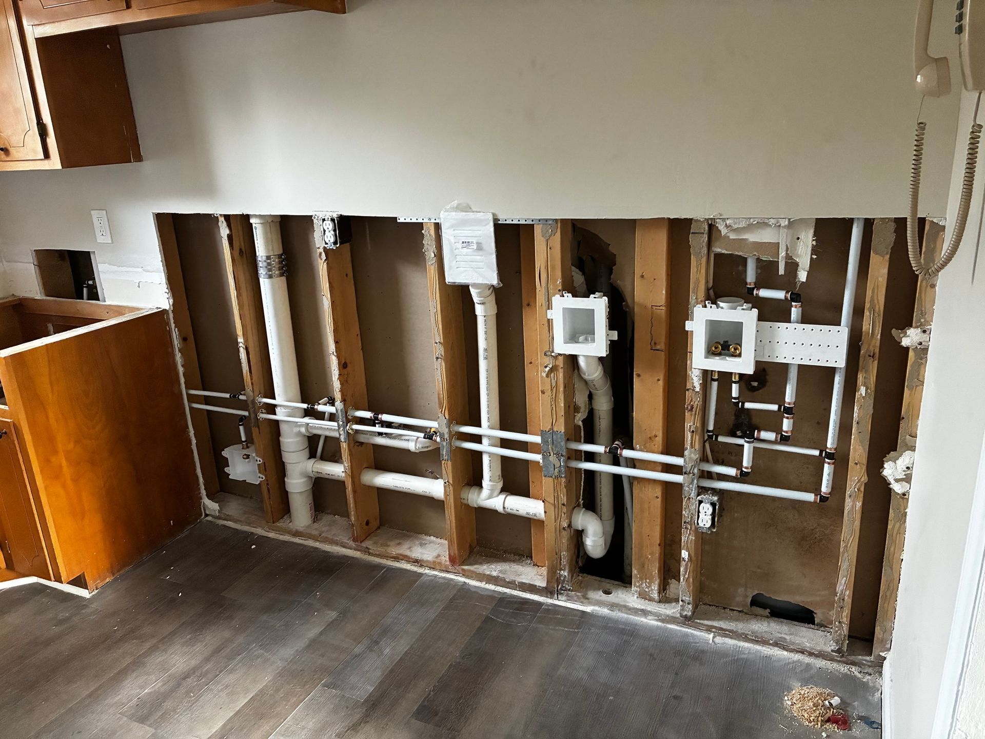 An unfinished interior wall showing exposed wooden studs, installed white plumbing pipes, and laundry connection boxes.