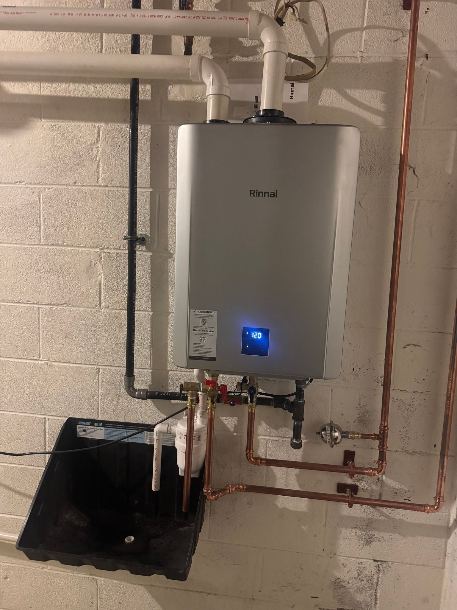A silver Noritz tankless water heater mounted on a white concrete wall with connected white PVC and copper pipes.