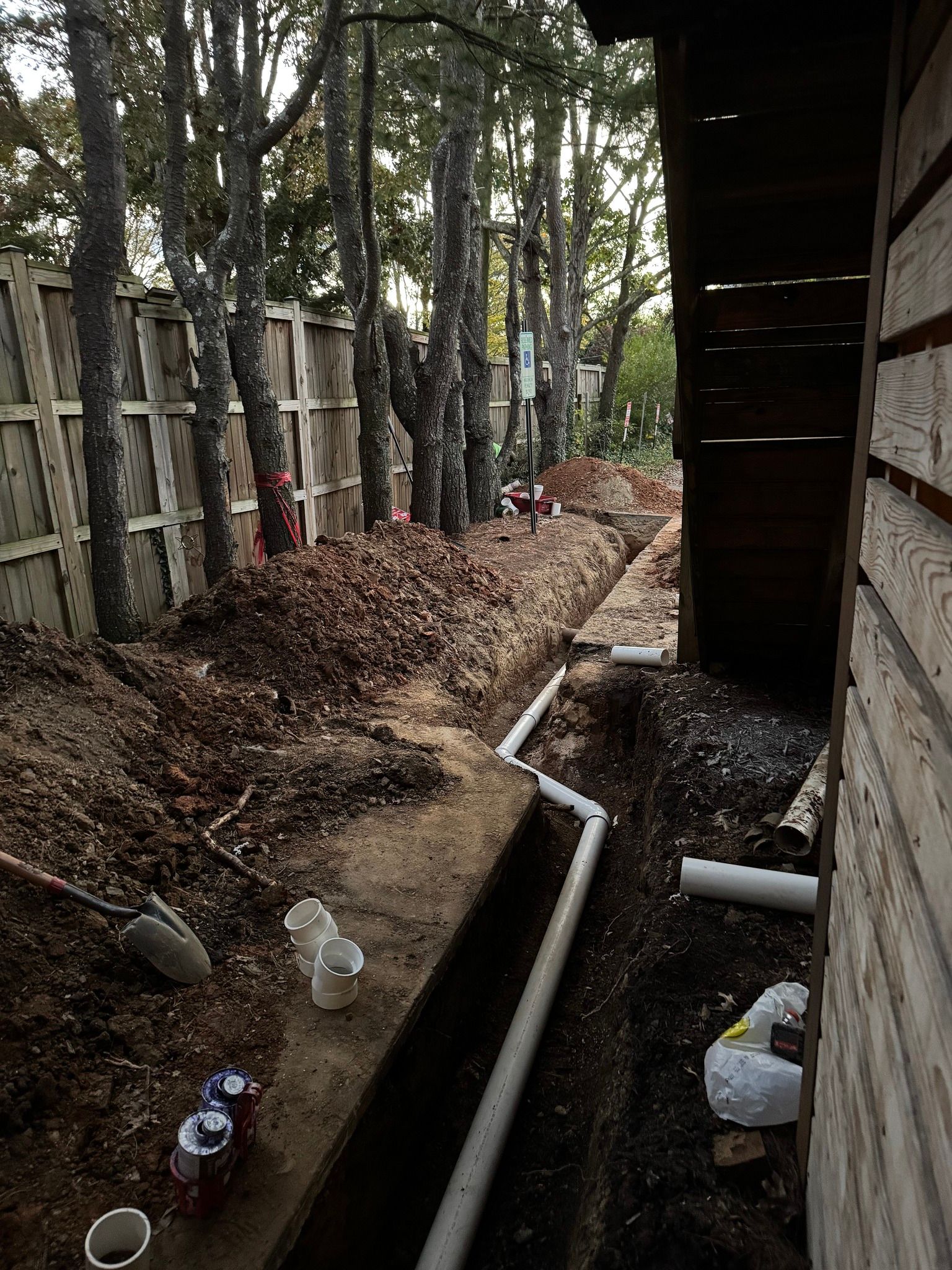 A trench containing a white PVC drain pipe runs along the side of a wooden building, with a fence and trees nearby.