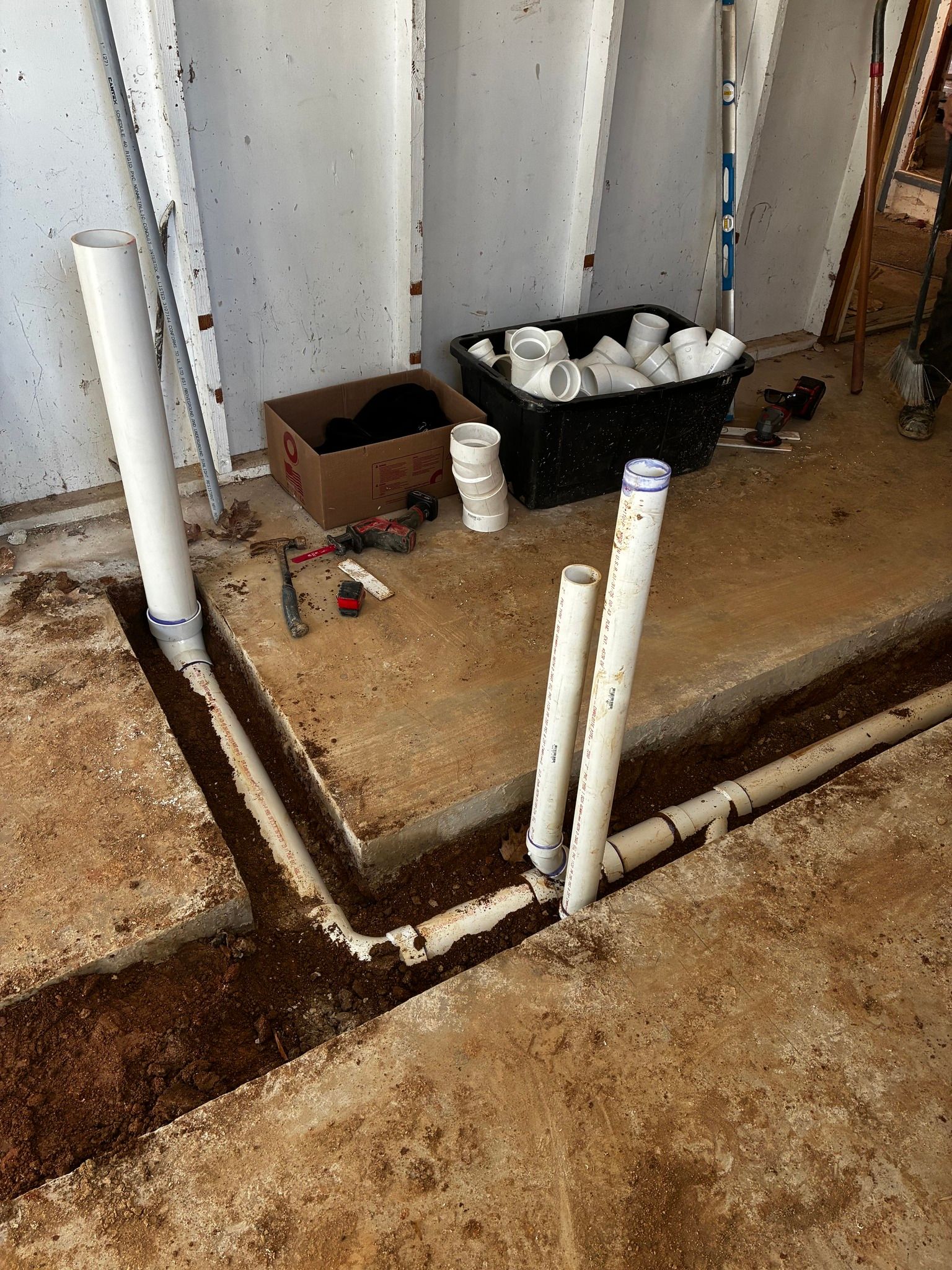 White PVC pipes protrude from trenches in a dirt floor near a concrete foundation, with construction materials nearby.