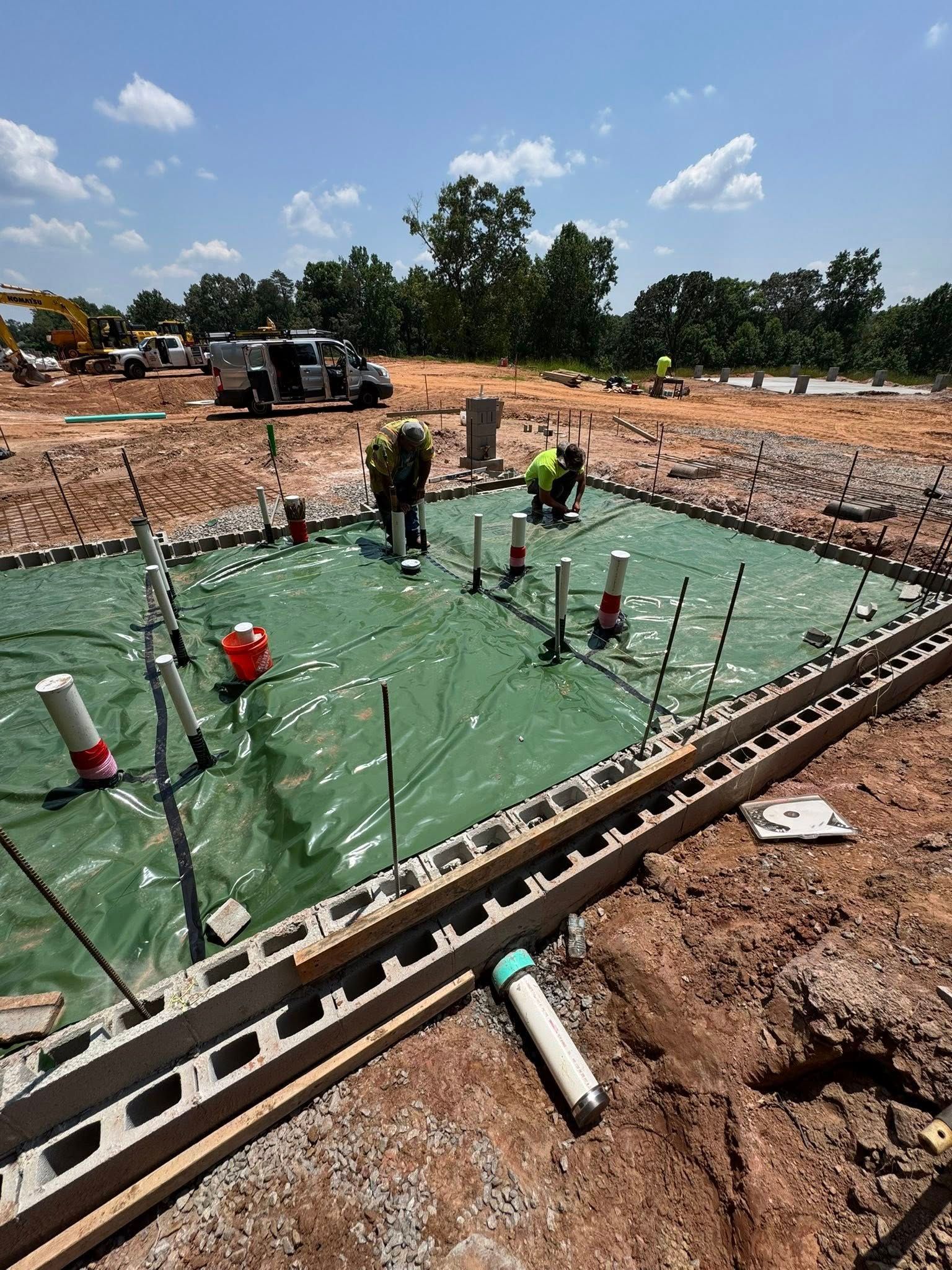 Two construction workers install plumbing pipes on a green vapor barrier inside a concrete block foundation at a site.
