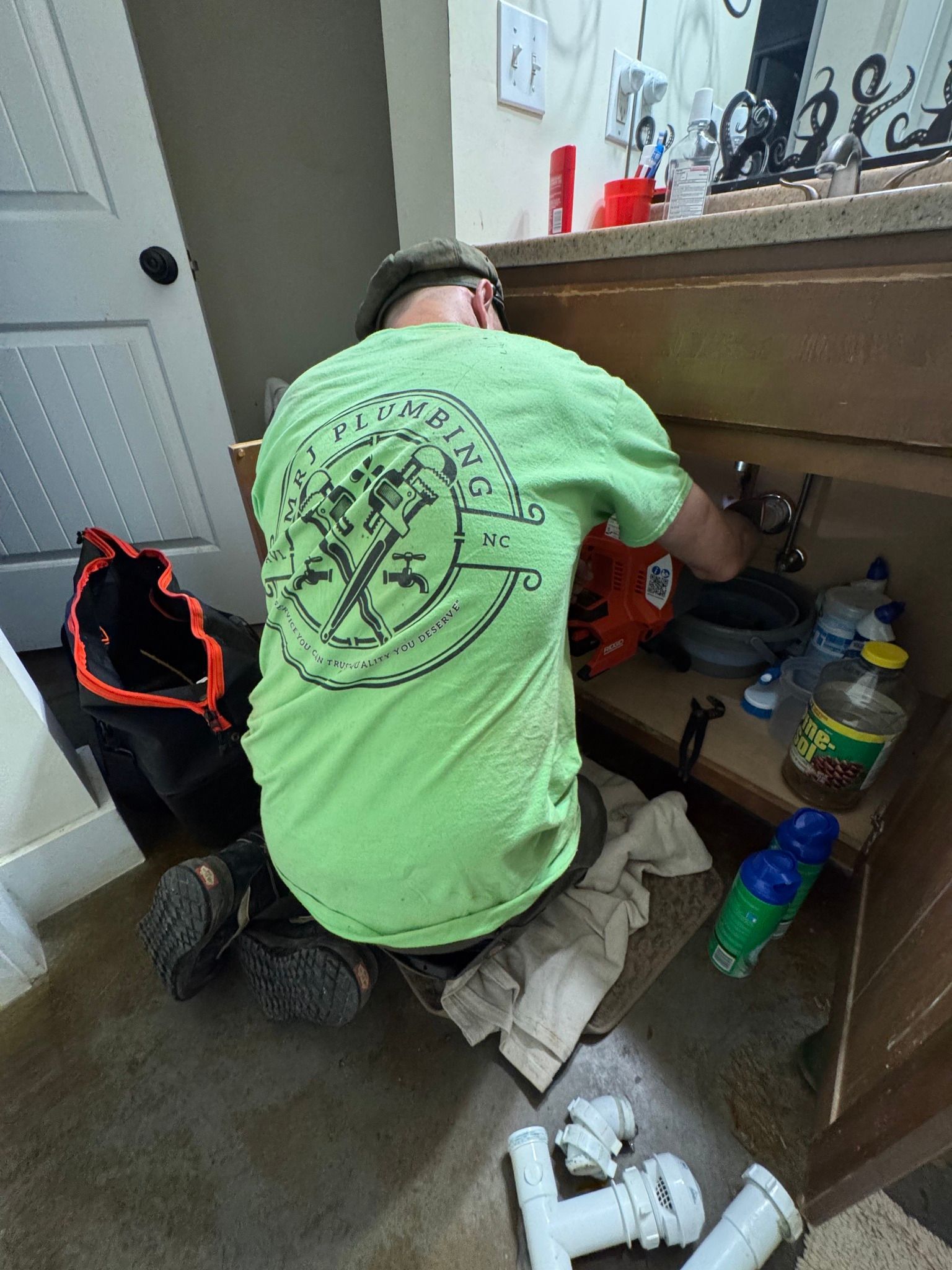 A person wearing a bright green t-shirt kneels on a bathroom floor, repairing pipes under a wooden sink cabinet.