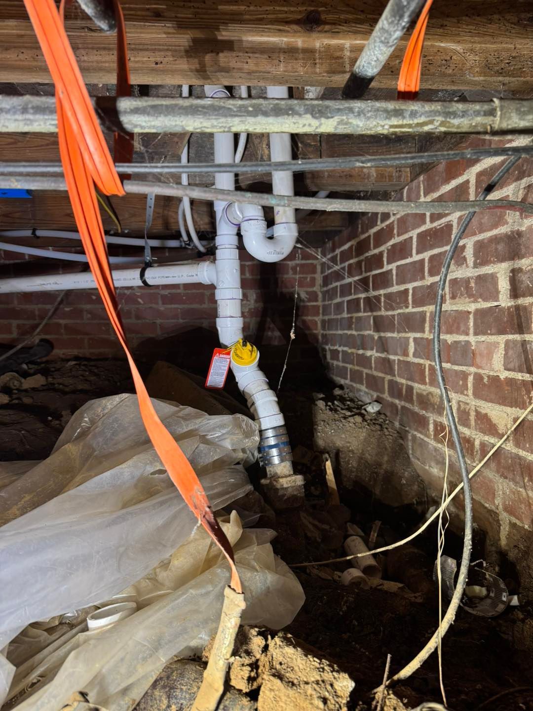 White PVC plumbing pipes with a yellow clean-out plug installed in a dark crawlspace with brick walls and debris.