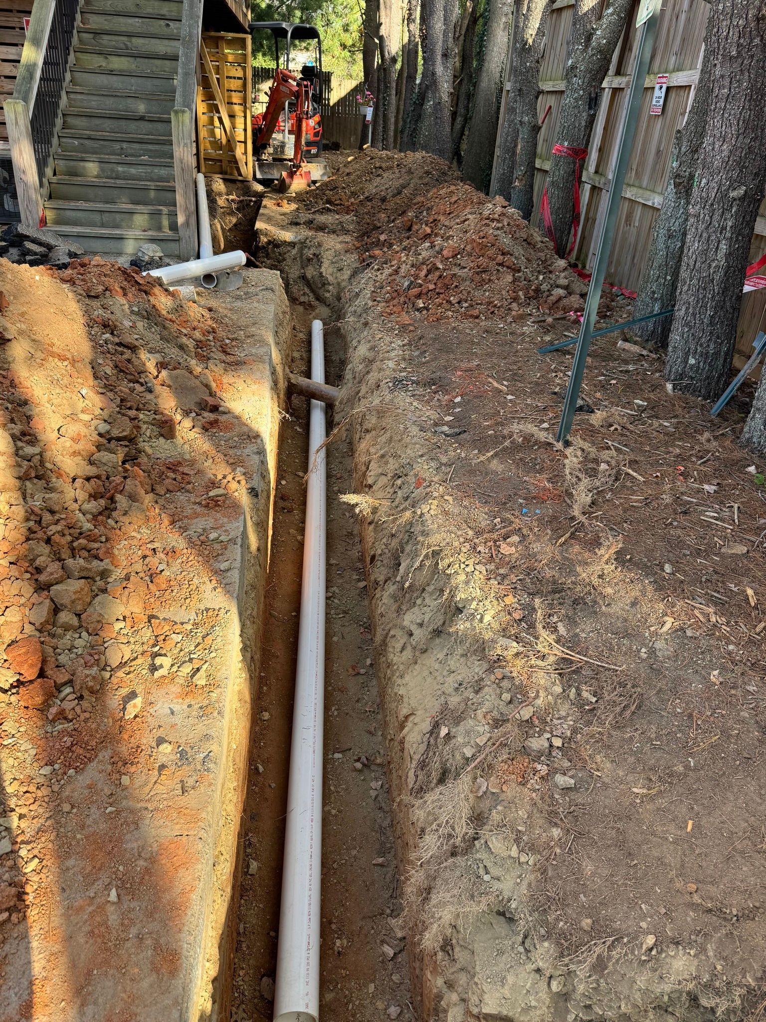A white PVC pipe lies in a narrow dirt trench excavated next to a wooden fence and stairs.