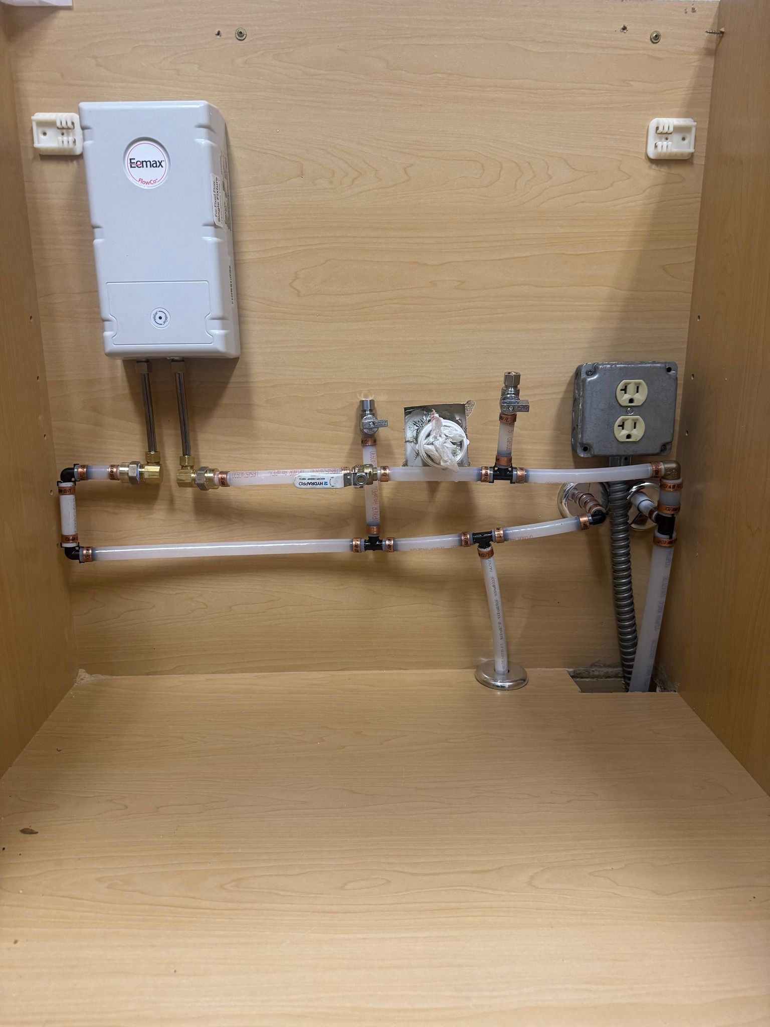 An under-cabinet view showing a white tankless water heater connected to white PEX piping and an electrical outlet box.