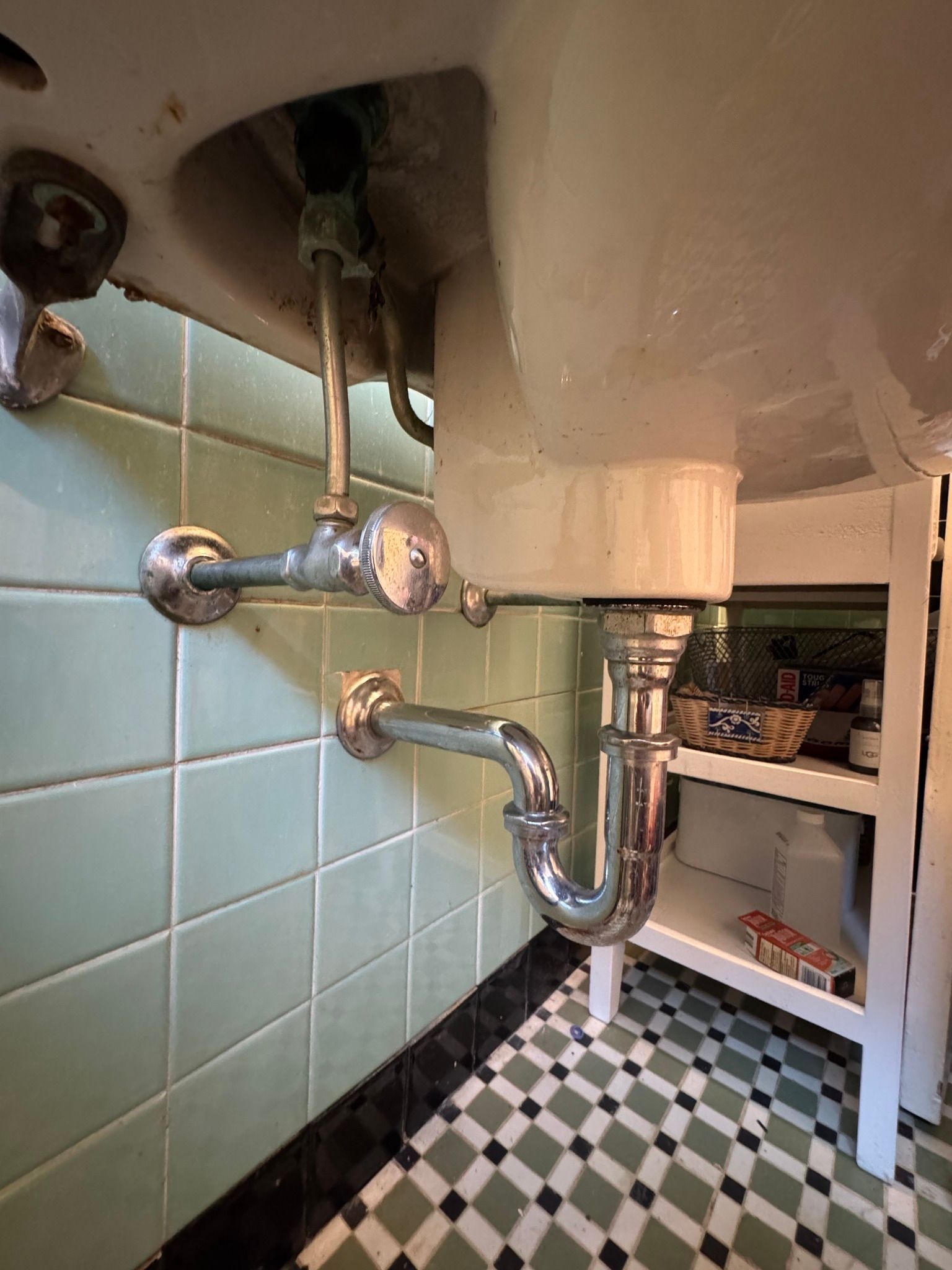 A wall-mounted bathroom sink with visible chrome plumbing pipes against green-tiled walls and a checkered floor.