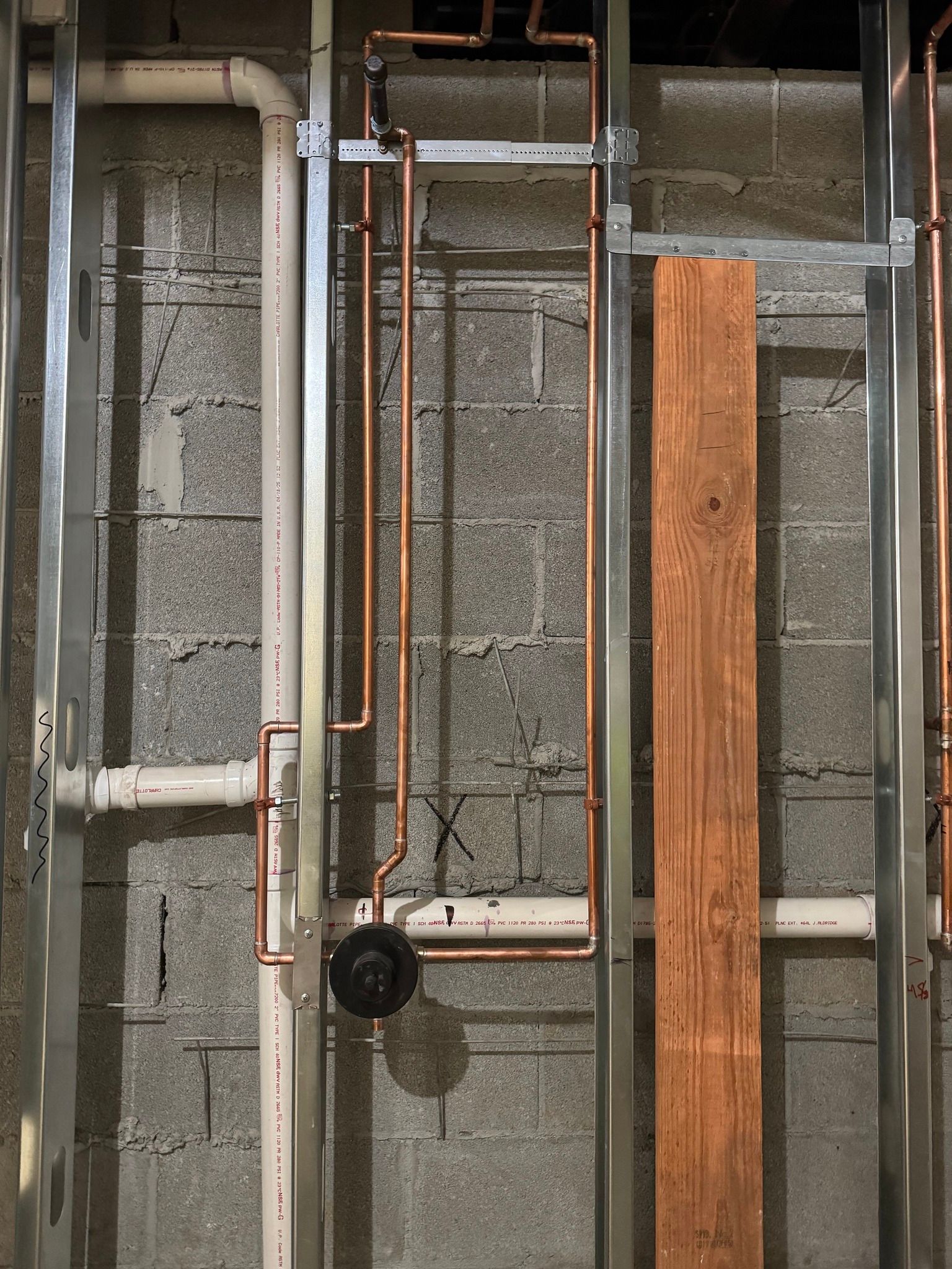 Copper plumbing pipes installed in a framed wall with a shower valve and white PVC pipes against a cinder block wall.