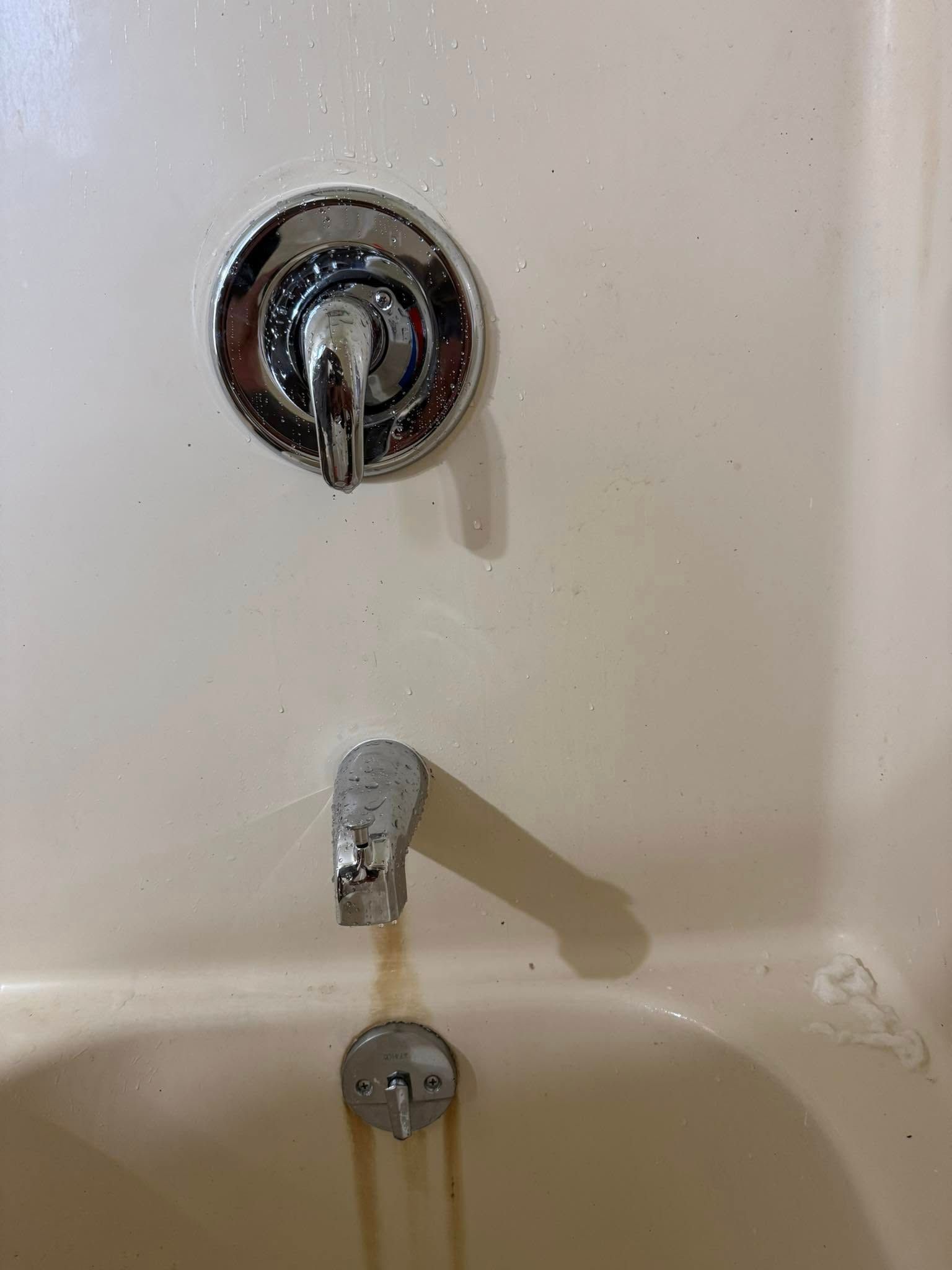 A bathtub faucet and shower handle on a beige wall, with orange water stains running down into the tub drain.