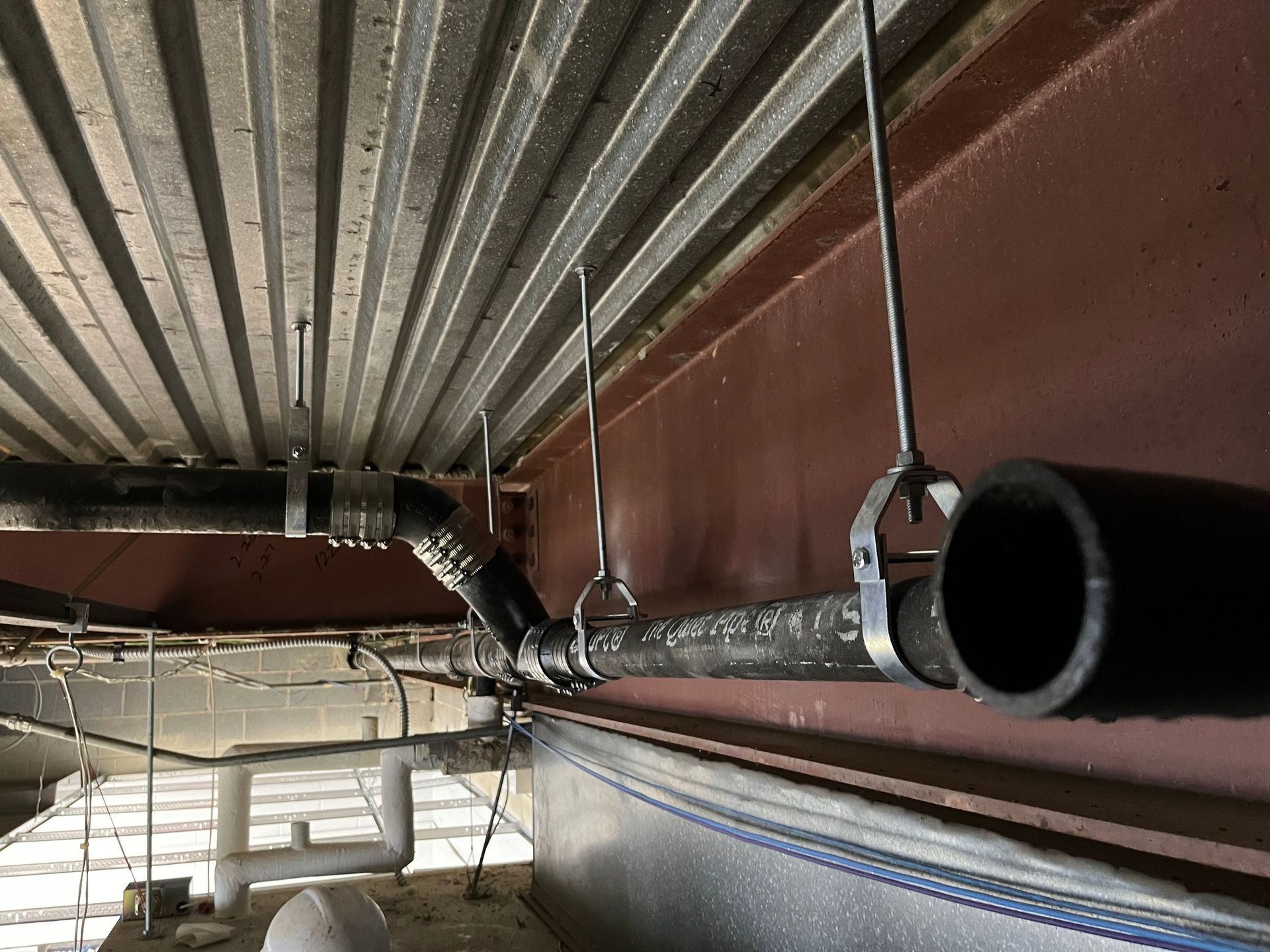 Industrial plumbing pipes suspended by metal rods from a corrugated metal ceiling and brown steel beam.